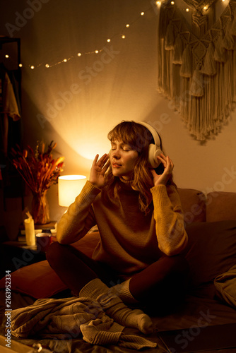 Young adult Caucasian woman sitting cross legged on bed wearing headphones with eyes closed, listening to music or meditating, representing mental wellbeing, vertical