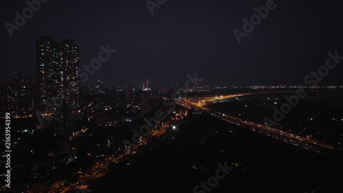An aerial view of Mumbai's high-rise buildings and beautiful nightlights. The sky is cloudy, and there is medium traffic on the road. The cityscape is filled with modern illuminated buildings.