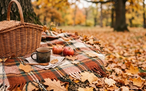 Autumn picnic setup with wicker basket warm drink and apples on a plaid blanket in a forest covered with fallen leaves representing cozy seasonal vibes and relaxation