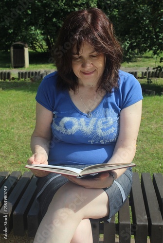 Smiling Mature Woman Reading a Book on a Wooden Park Bench in Summer Sunlight.

