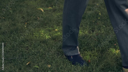 Various video clips of a bearded man wearing sunglasses and a polo shirt walking in a green park. He strolls along a paved path and adjusts his sleeve under natural daylight among trees.
