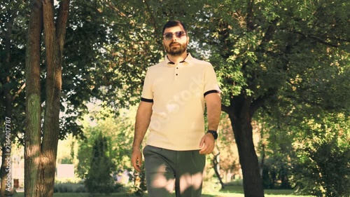 Various video clips of a bearded man wearing sunglasses and a polo shirt walking in a green park. He strolls along a paved path and adjusts his sleeve under natural daylight among trees.