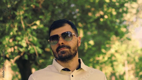 Various video clips of a bearded man wearing sunglasses and a polo shirt walking in a green park. He strolls along a paved path and adjusts his sleeve under natural daylight among trees.