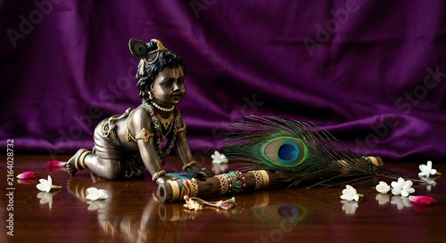 Laddu Gopal statue with peacock feather, flute, and white jasmine flowers on purple fabric.