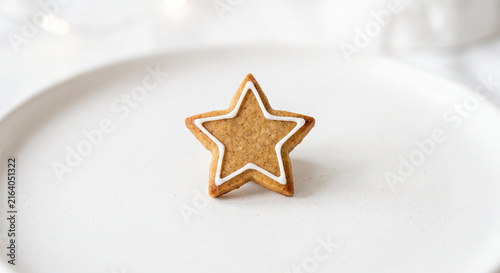 A single star shaped gingerbread cookie with white icing on a plain white plate with bokeh lights in the background horizontal