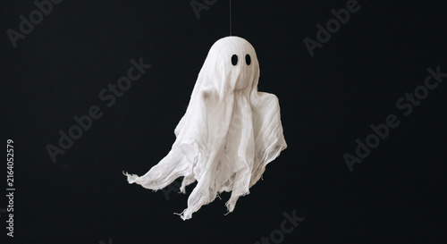 A small white ghost decoration with black eyes hanging against a dark black background creating a spooky Halloween atmosphere