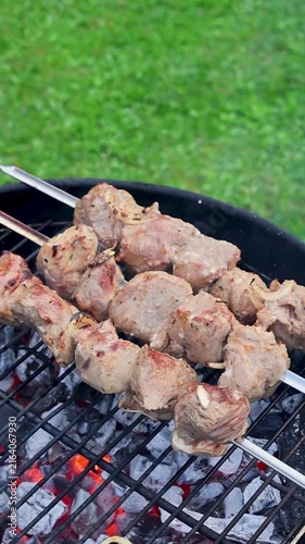 Marinated pieces of meat grilling on metal skewers over a hot charcoal grill in a summer garden with fresh green grass in the background. Close-up barbecue scene with smoky grilled meat