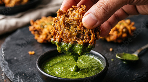 Close Up View of Human Hand Dipping Crispy Golden Onion Pakora into Fresh Green Mint Chutney with Drip on Dark Slate Board