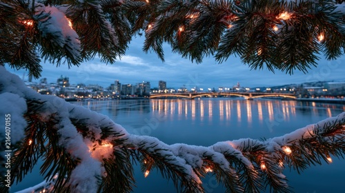 Snow covered pine branches with fairy lights frame a city skyline and bridge reflection at dusk