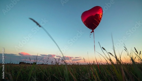 Red heart balloon floating above a grassy meadow at sunset,