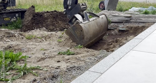 Excavator bucket hovers over a dirt mound at a construction site, with grass and gravel in the foreground 