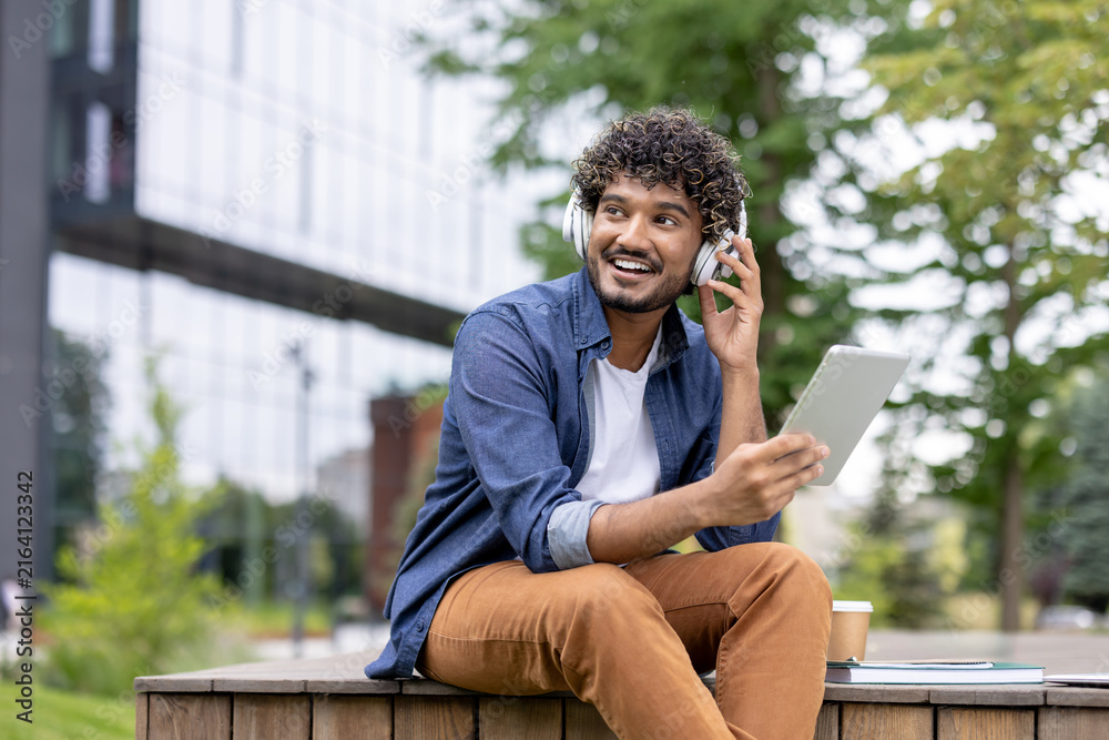 Fototapeta premium Young man with curly hair enjoying music through headphones, holding a digital tablet, and smiling while relaxing on a wooden bench in an urban park