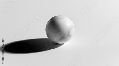 A marble ball sits on a plain white surface. The ball is smooth with a subtle pattern. A shadow is cast on the surface, creating contrast