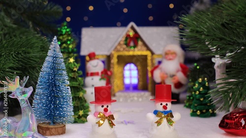Two snowmen in front of Christmas trees and a house with Santa Claus