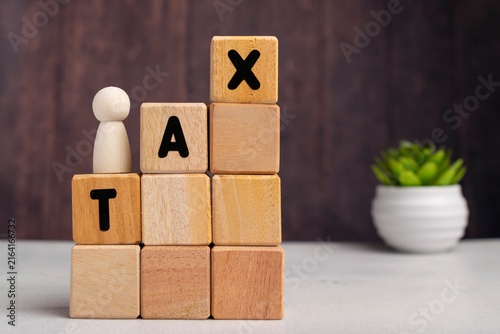 Obraz na płótnie Wooden block tower spelling TAX diagonally with a small wooden peg figure standing on top, financial growth concept on dark background