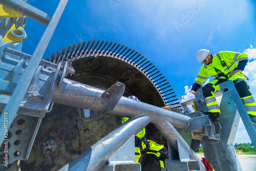 Wind farm technician at ground level reaches up past the galvanised cradle tubes toward his colleague on the frame, the pair working together at the blade root end