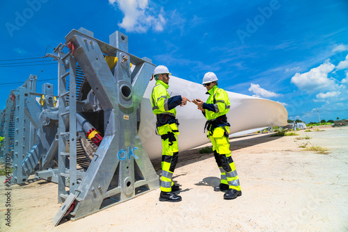 Engineer gestures toward the trailing edge of a wind turbine blade while his colleague listens, the pair standing on hot concrete beside the galvanised root cradle