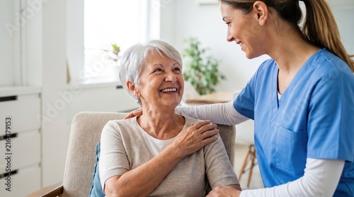 Smiling senior woman sitting on sofa, holding shoulder as a female caregiver in blue uniform supports her; warm home care moment in a bright room