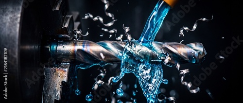 High speed drill bit boring through steel component with blue cooling fluid splashing and metal shavings flying in cnc machine process