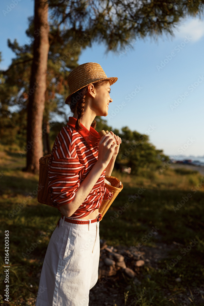 Naklejka premium Young woman enjoying lifestyle at sunset in nature. She wears a straw hat and striped shirt. Concept of summer vacation, travel, relaxation, leisure, and outdoor hobby.
