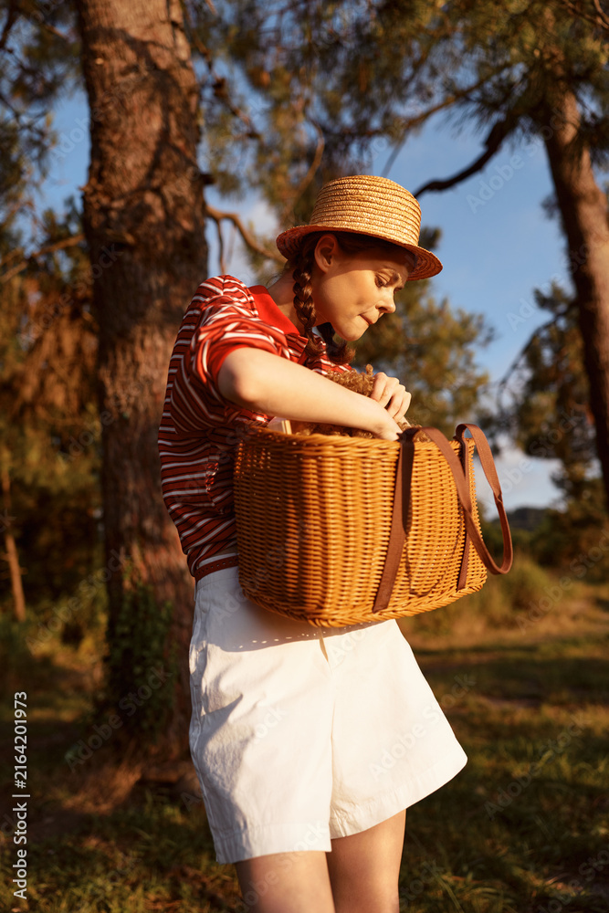 Naklejka premium Young woman outdoors in a straw hat and striped shirt holds a wicker basket, smiling softly as she enjoys a sunny countryside moment. Soft glow highlights leisure.