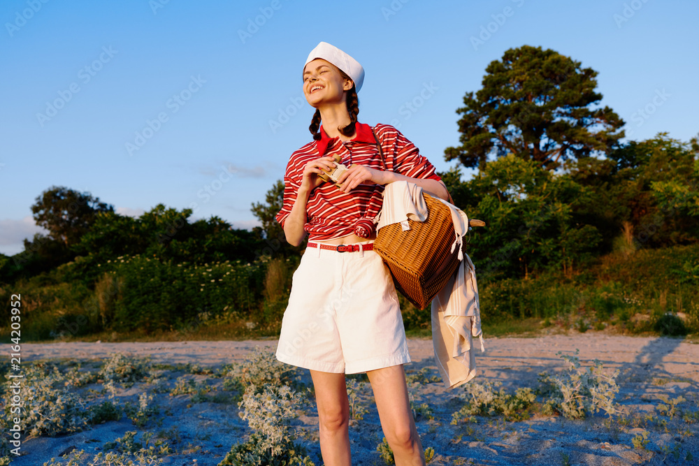 Naklejka premium Young woman enjoying lifestyle at picnic in nature. She wears retro outfit with hat and carries wicker basket. Sunny outdoor setting creates happy relaxed summer mood.