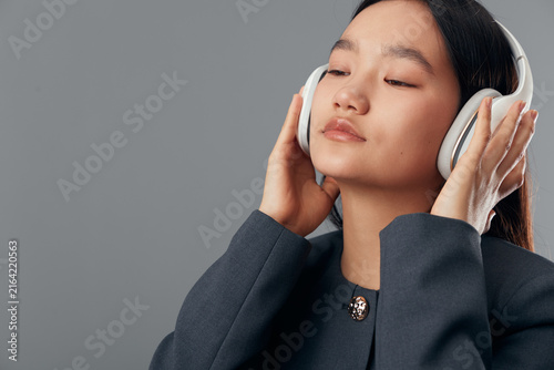 Obraz na plátne Young woman with closed eyes wearing white headphones and dark jacket listening to music or audio content in calm, relaxed pose on neutral gray background in studio