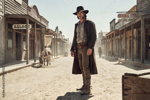 Cowboy in a wide brim hat and dark coat standing on the dusty main street of a vintage western town with wooden buildings and a donkey nearby