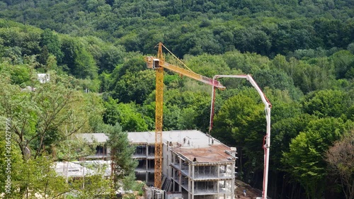 A high-angle view of workers in safety vests operating a yellow tower crane and a concrete pump truck with a long distributor boom, pouring mixture into formwork on a multi-story building near forest.