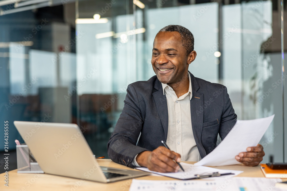 Obraz premium Experienced entrepreneur smiling while reviewing financial paperwork and signing contracts at his desk, managing business operations with a laptop in a modern corporate workspace