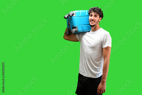 Young man carrying a blue suitcase on his shoulder, smiling and looking at the camera against a bright green background, representing travel, holidays, vacation planning and tourism.