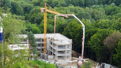 A high-angle view of a yellow tower crane and a concrete pump truck working near a forest, as safety-vested workers control a hose to pour mixture into formwork on a multi-story building.