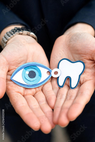 Two open hands hold layered paper cut symbols of a blue eye and a white tooth, a clean conceptual still life image representing vision and dental health care coverage.