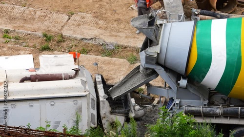 A concrete mixer truck discharges fresh liquid cement through a chute into a white construction pump machine on a dusty daytime building site for a real estate project.