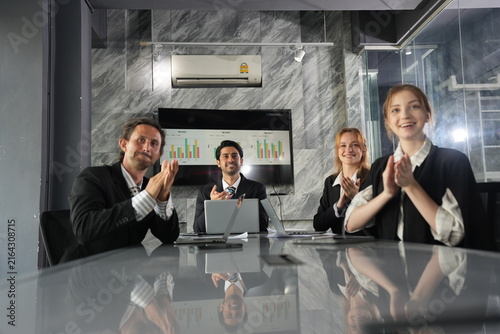 Canvastavla Wide angle of a cheering four person office team applauding behind a laptop strewn conference table, every clap doubled by reflections on its polished silver surface