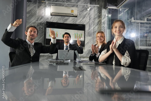 Fists pumped high by two cheering businessmen while smiling female colleagues keep the applause going, pure elation around the glossy conference table at night in a bright modern coworking.