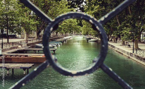 Konstfotografi Vue sur un canal bordé d'arbres et de bateaux à travers le motif circulaire d'une grille en fer forgé à Annecy