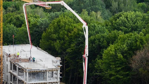A high-angle view of a yellow tower crane and concrete pump truck working near a forest, as safety-vested workers control a hose to pour mixture into formwork on a multi-story building.