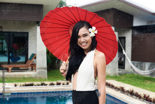 Portrait of smiling woman with flower in her hair holding a red tradtional umbrella at a swimming pool