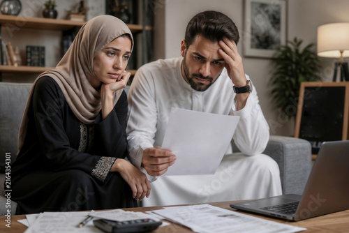 A Muslim couple sits tensely, reviewing their accounts together. They have large outstanding balances at the bank. The man holds his head, clearly tense. Bad news