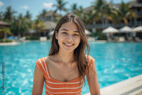 Sunlit poolside with a smiling young woman. She wears an orange-striped top, hair gently tousled. Tropical palms frame a calm, blue swimming pool