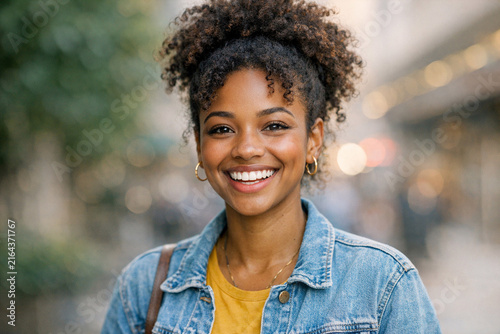 Young woman with curly hair and gold hoop earrings smiling at camera with joy. She is wearing a denim jacket over yellow shirt, set against city street background.