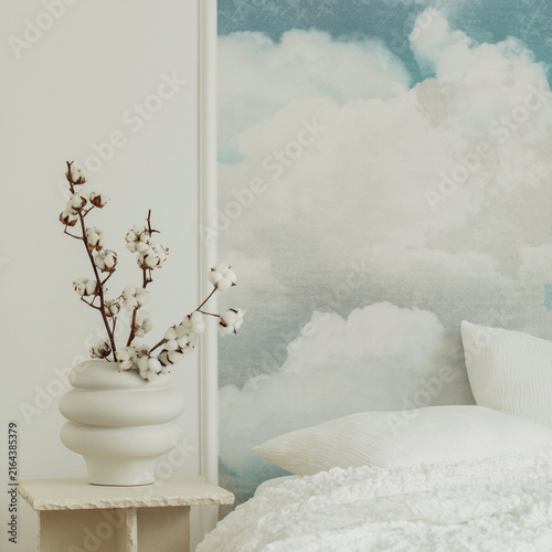 Minimalist bedroom interior design with a cloudy sky wall mural, white ceramic vase with dried cotton branch, and cozy white bed linen