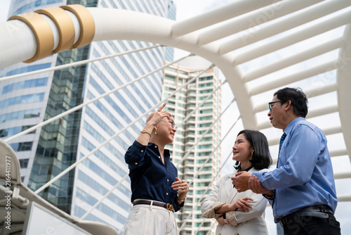 Three business professionals having discussion on outdoor pedestrian skywalk bridge with modern city skyscrapers background, corporate teamwork conversation