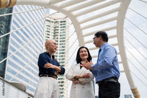 Three business professionals having discussion on outdoor pedestrian skywalk bridge with modern city skyscrapers background, corporate teamwork conversation