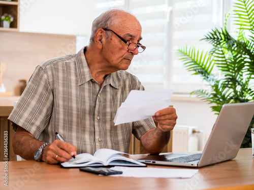 Senior man writing notes while using laptop for electronic banking, checking expenses and reviewing his personal financial information.