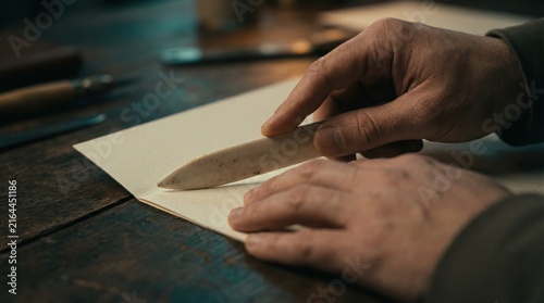 A hand presses a tool against paper to make a crease in a workshop during evening hours. Tools and materials are scattered around