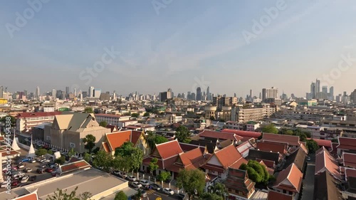 Wide nighttime panoramic view of Bangkok from a Buddhist temple observation deck showing temple grounds, residential roofs, and distant skyscrapers at sunset.