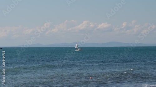 a white sailboat cruising on a deep blue sea with rolling waves and a small motorboat briefly appears speeding on the left.