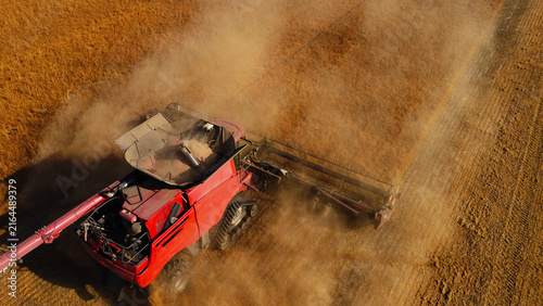 Foto A high-angle top-down aerial drone photograph capturing a large red combine harvester kicking up a thick cloud of golden dust as it operates across a harvested grain field